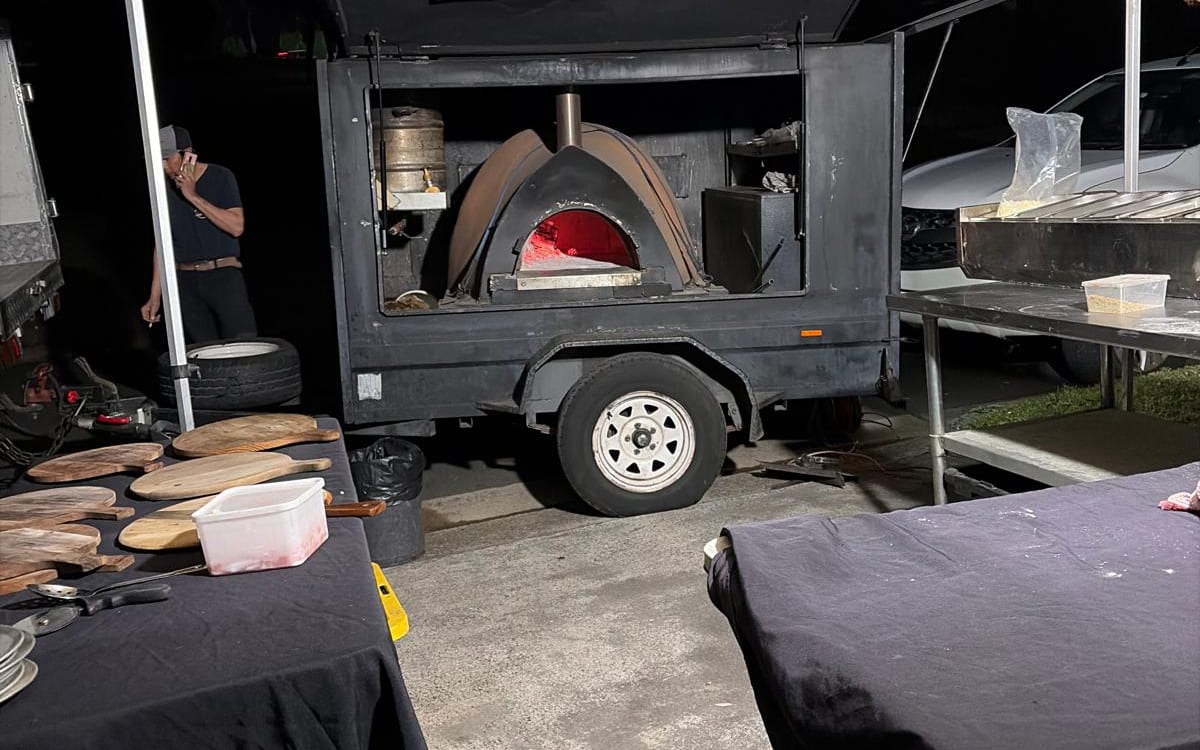 The wood-fired oven glowing inside the trailer at a night-time community event, pizza boards ready